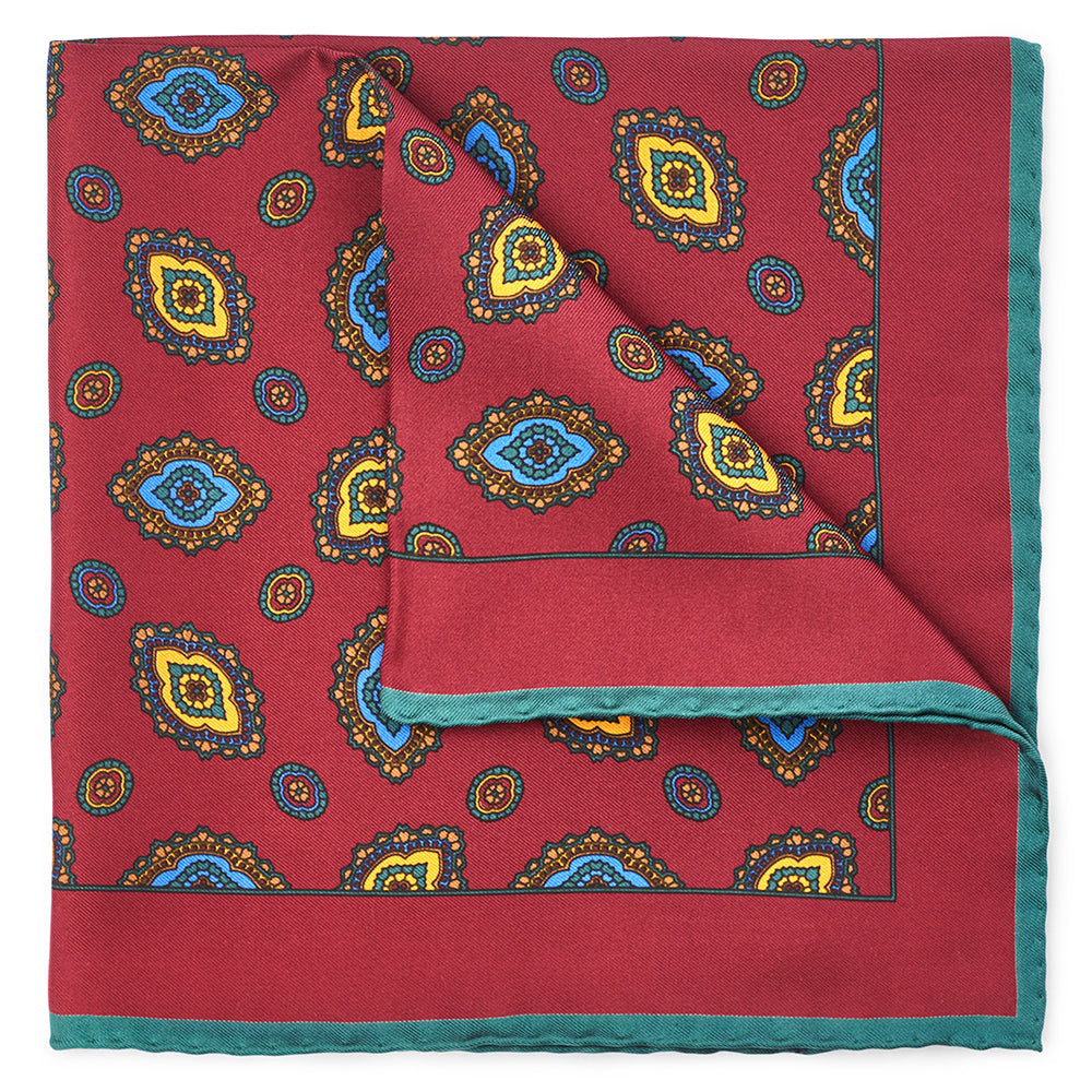 Red silk pocket square with an ornate diamond print in blue, yellow and green, with a green border